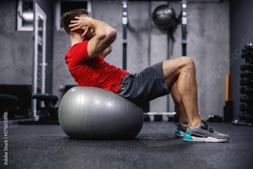 Crunches on Pilates or yoga balls. A muscular and handsome male person in sportswear performs sit-ups on a fitness ball in a modernly equipped dark atmosphere of the sports center