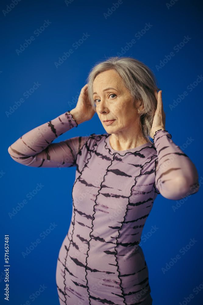 senior woman in purple dress adjusting grey hair while posing isolated on blue senior woman in purple dress adjusting grey hair while posing isolated on blue