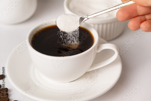 The process of pouring white sugar from a spoon into a white cup of coffee. Sugar addict, diabetes.
