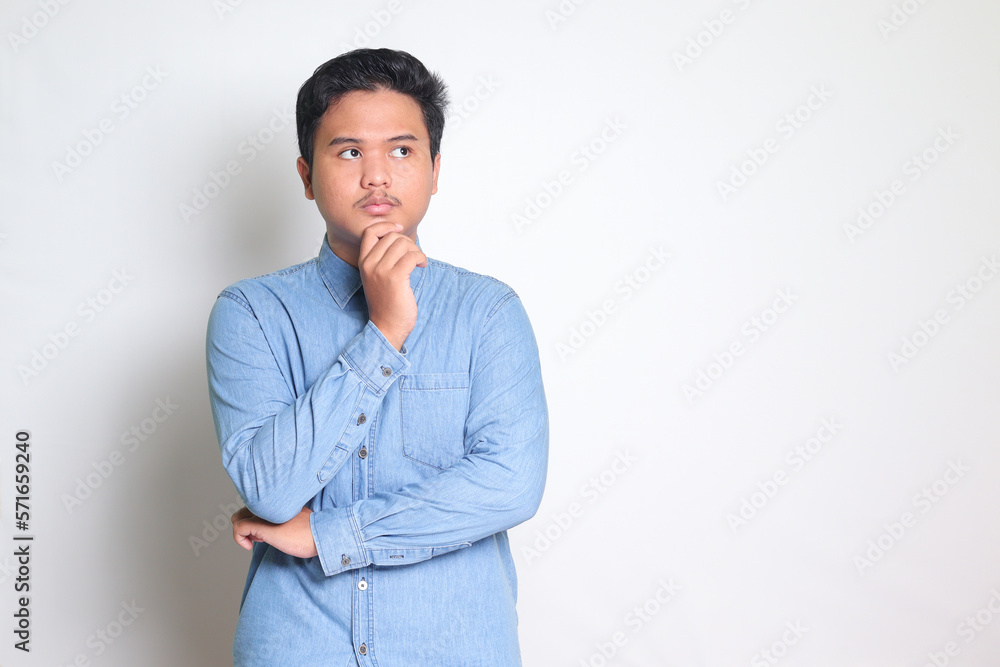 Portrait of confused Asian man in blue shirt thinking about question with hand on chin. Isolated image on white background Portrait of confused Asian man in blue shirt thinking about question with hand on chin. Isolated image on white background