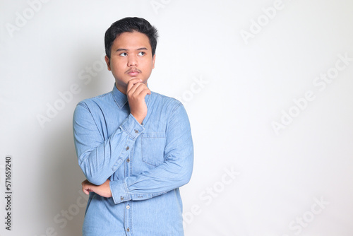 Portrait of confused Asian man in blue shirt thinking about question with hand on chin. Isolated image on white background