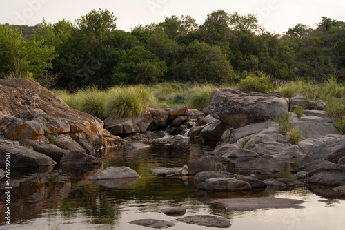 View of the calm river flowing across the rocky hill and forest at sunset. 