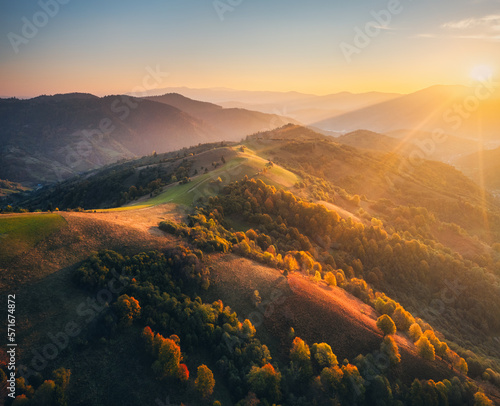 Aerial view of beautiful hills and mountains in fog at sunset in autumn in Ukraine. Colorful landscape with foggy woods, meadows, golden sunlight. Orange trees. Forest in fall. Drone view. Nature