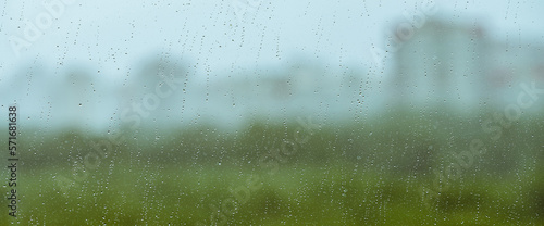 Atmospheric minimal backdrop with rain droplets on glass. Wet window with rainy drops and dirt spots closeup. Blurry buildings and green trees under blue sky against dirty window glass with raindrops.