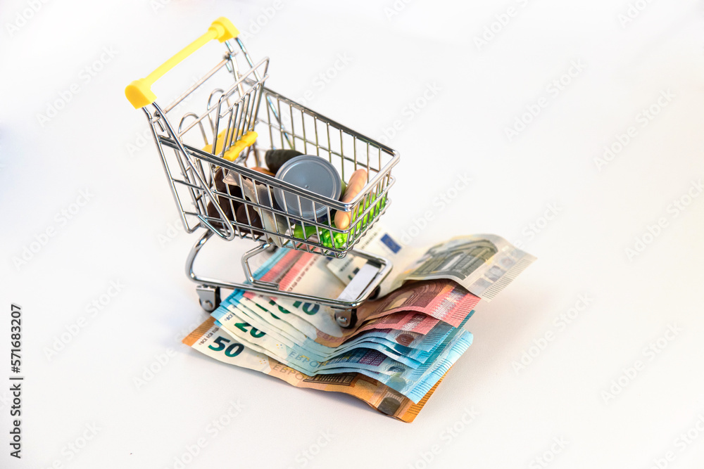 Supermarket shopping cart with essential goods and several euro banknotes on a white background. Conceptual image about purchasing power, family expenses, essential goods,consumer society. Copy space