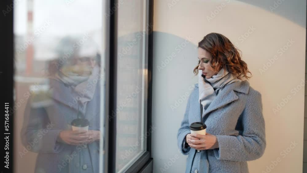 An elegant middle-aged woman stands at the window of a building in outerwear An elegant middle-aged woman stands at the window of a building in outerwear