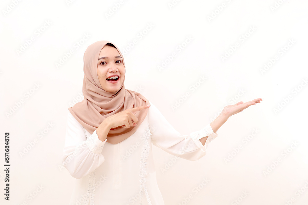 Portrait of amazed young asian muslim woman standing while presenting and pointing something on blank space