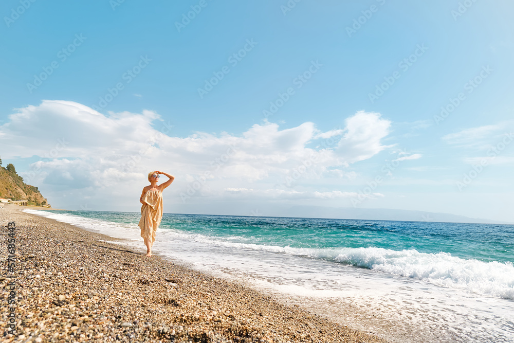 Pretty blond woman walking on pebble beach enjoying sunny windy day near blue sea. Travel concept. Pastel muted color.