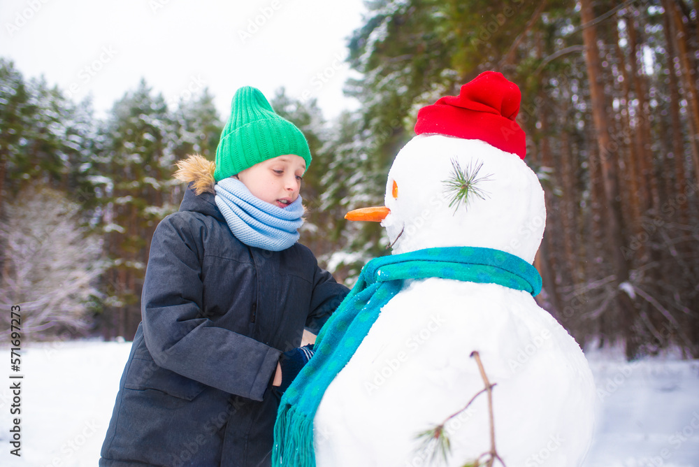 the boy made a snowman, a happy child plays in a snowy winter forest, straightens a scarf and a hat for a snowman