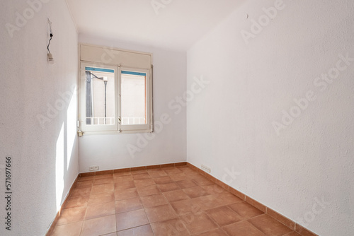 An empty spacious room with old square worn caramel colored floor tiles and window with natural sun light, white faded walls and ceiling. The wires for the lamp stick out in the wall. © Pavel