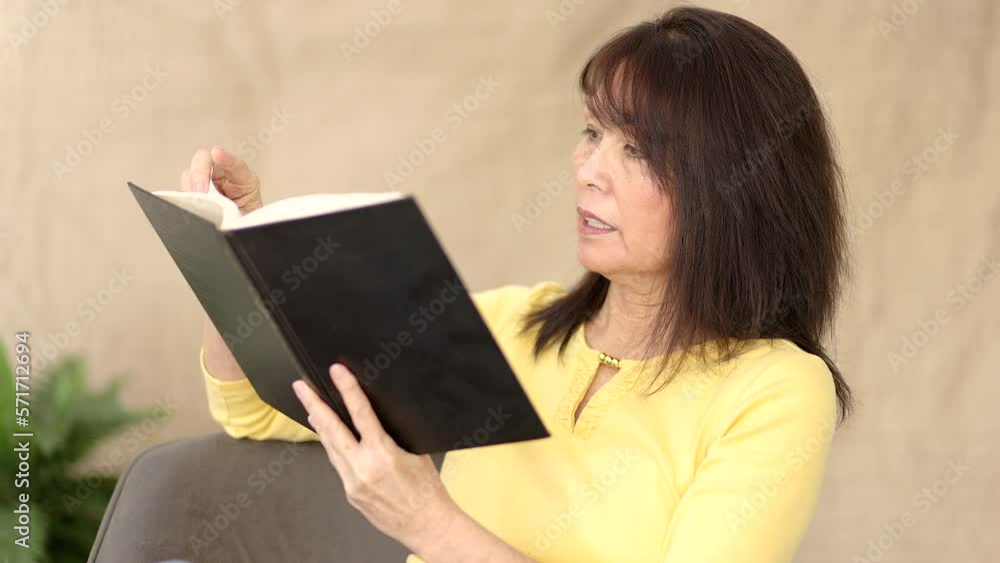 Attractive asian woman in yellow blouse enjoying reading book sitting in comfortable living room chair at home Attractive asian woman in yellow blouse enjoying reading book sitting in comfortable living room chair at home