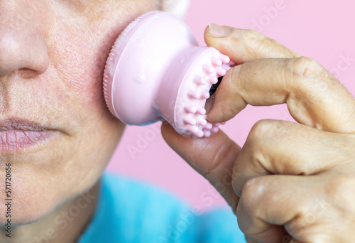 senior old granny lady elderly woman using silicone sponge for face cleaning,vacuum cup for massage.smiling female showing wrinkle undereye bags on forehead.isolated woman on pink wearing headband photo