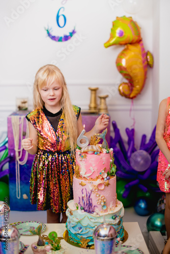 Indoor shot of a rather joyful little girl with blond hair making a wish before blowing out the candle. Celebrates her 6th birthday in a trendy dress, with a huge nautical-themed cake. Happy childhood photo