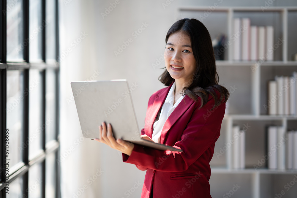 Fototapeta premium Asian businesswoman standing in the office room near the window while holding laptop computer in her hands.