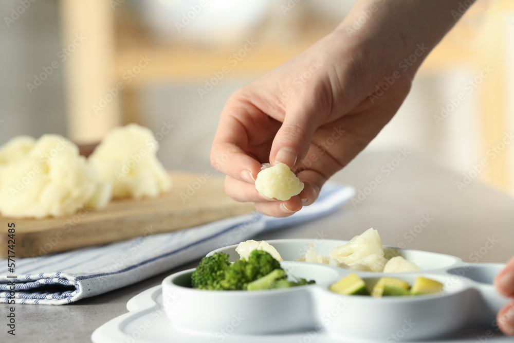 Baby food. Woman putting piece of cauliflower into section plate with different vegetables at grey textured table, closeup Baby food. Woman putting piece of cauliflower into section plate with different vegetables at grey textured table, closeup