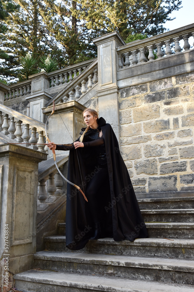Portrait of beautiful female model with blonde plait, wearing black leather catsuit and hooded cloak, fantasy assassin warrior. Standing pose holding archery bow and arrow weapon, castle background.