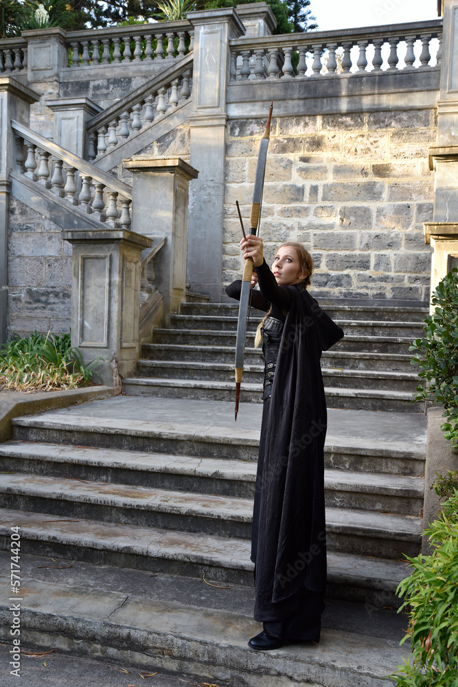 Portrait of beautiful female model with blonde plait, wearing black leather catsuit and hooded cloak, fantasy assassin warrior. Standing pose holding archery bow and arrow weapon, castle background. Portrait of beautiful female model with blonde plait, wearing black leather catsuit and hooded cloak, fantasy assassin warrior. Standing pose holding archery bow and arrow weapon, castle background.