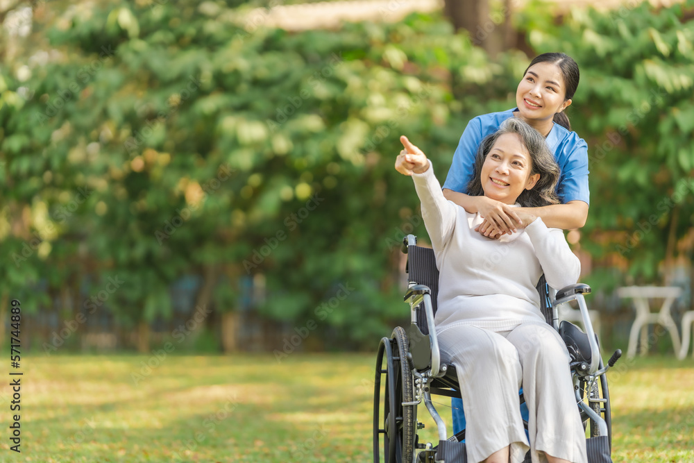 Young asian care helper with asia elderly woman on wheelchair relax together park outdoors to help and encourage and rest your mind with green nature. Hand pointing forward Young asian care helper with asia elderly woman on wheelchair relax together park outdoors to help and encourage and rest your mind with green nature. Hand pointing forward
