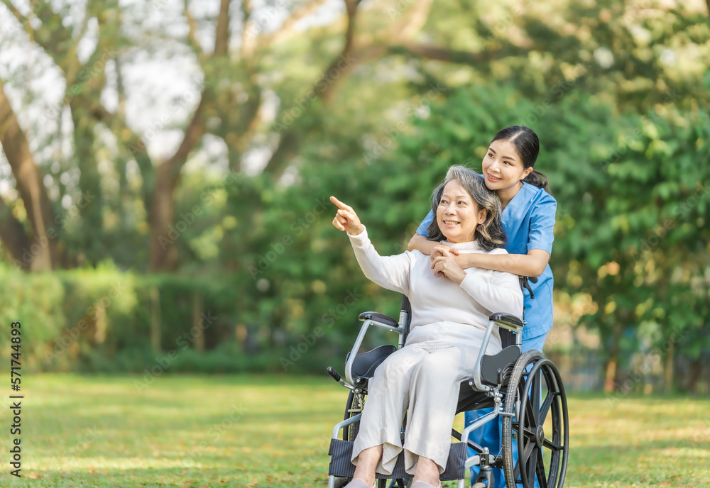 Young asian care helper with asia elderly woman on wheelchair relax together park outdoors to help and encourage and rest your mind with green nature. Hand pointing forward Young asian care helper with asia elderly woman on wheelchair relax together park outdoors to help and encourage and rest your mind with green nature. Hand pointing forward