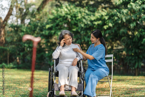 Young asian care helper with asia elderly woman on wheelchair relax together park outdoors to help and encourage and rest your mind with green nature. Use a stethoscope to listen to your heartbeat.