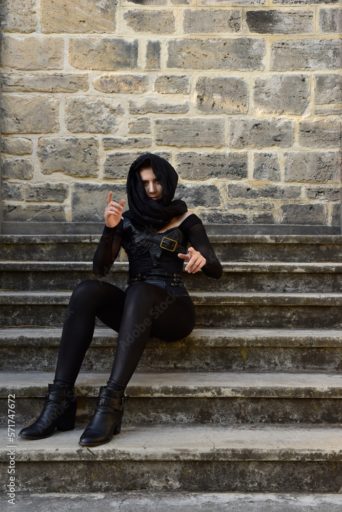 Portrait of beautiful female model with blonde plait, wearing black leather catsuit and flowing hooded cloak, fantasy assassin warrior. Posing in castle background with stone staircase. Portrait of beautiful female model with blonde plait, wearing black leather catsuit and flowing hooded cloak, fantasy assassin warrior. Posing in castle background with stone staircase.