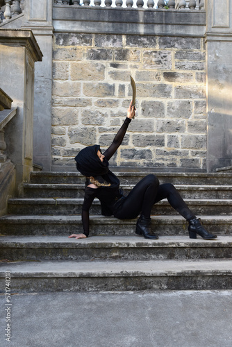 Portrait of beautiful female model with blonde plait, wearing black leather catsuit and flowing hooded cloak, fantasy assassin warrior. Posing in castle background with stone staircase.