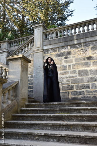 Portrait of beautiful female model with blonde plait, wearing black leather catsuit and flowing hooded cloak, fantasy assassin warrior. Posing in castle background with stone staircase.