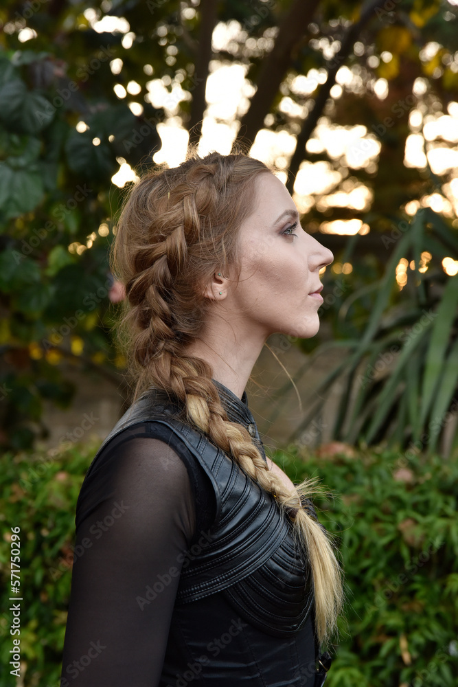 Close up portrait of beautiful female model with blonde plait, wearing black leather catsuit, fantasy assassin warrior. Posing in forest tree background Close up portrait of beautiful female model with blonde plait, wearing black leather catsuit, fantasy assassin warrior. Posing in forest tree background
