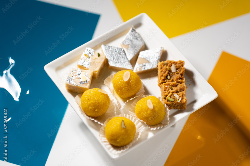 A plate of traditional Indian desserts — laddu, mawa burfi, and mohanthal — against a colorful background.
