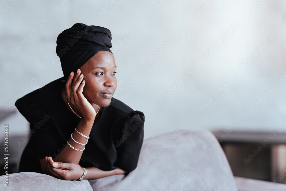 Pensive African girl in black turban and traditional dress looks aside leaning on couch against wall with empty space. Thoughtful young adult woman relaxing home. Muslim female indoors.