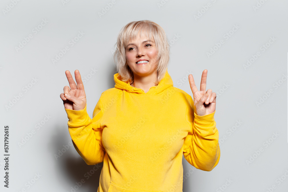 Studio portrait of smiling blonde woman in yellow hoodie sweater, shows peace gesture, on white background. Studio portrait of smiling blonde woman in yellow hoodie sweater, shows peace gesture, on white background.
