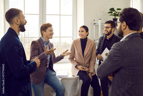 Group of a young happy smiling business people chatting after a meeting. Cowokers listening to their colleague and laughing. Company employees talking in the office in an informal setting. photo