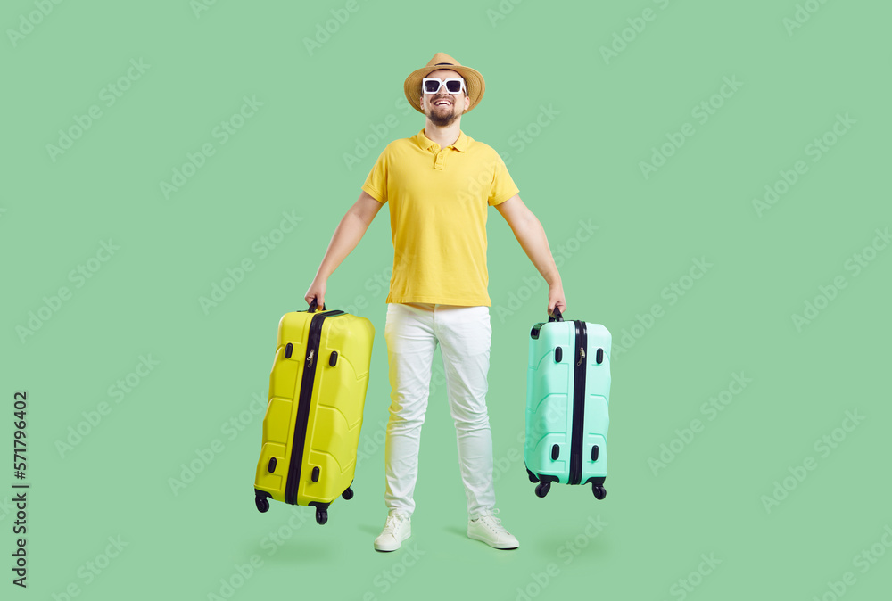 Happy young man in summer hat and sunglasses isolated on green studio background hold suitcases ready for vacation. Smiling male excited with holiday or trip. Summertime adventure concept. Happy young man in summer hat and sunglasses isolated on green studio background hold suitcases ready for vacation. Smiling male excited with holiday or trip. Summertime adventure concept.