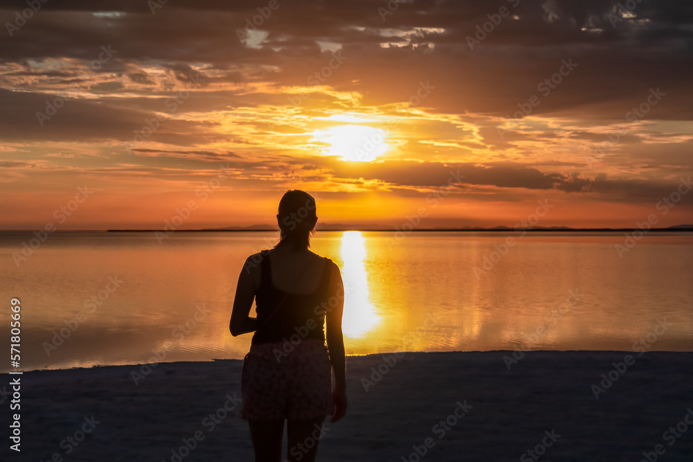 Silhouette of woman watching stunning sunrise of lake Bonneville Salt Flats, Wendover, Western Utah, USA, America. Dreamy red colored clouds mirroring on the water surface creating romantic atmosphere