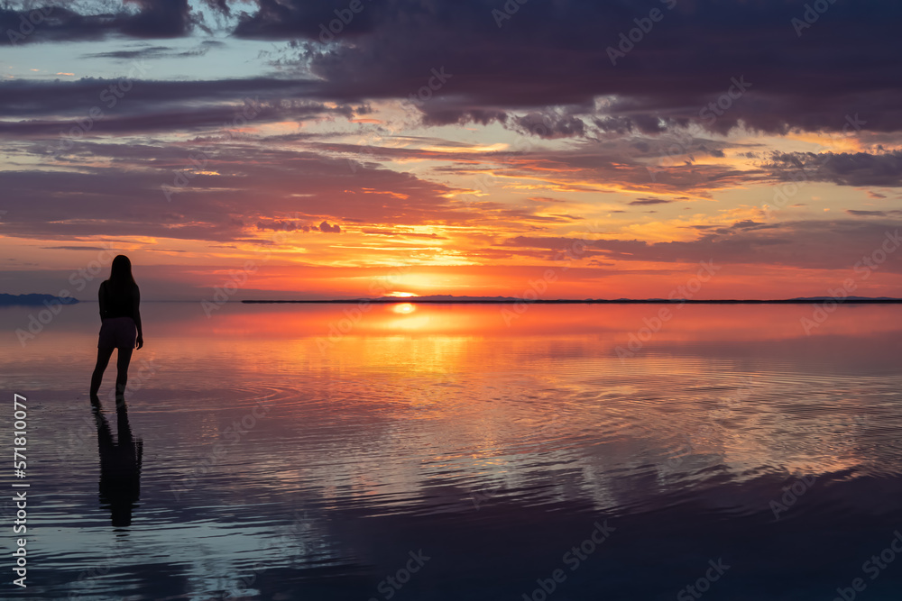 Silhouette of woman walking into the sunrise of lake Bonneville Salt Flats, Wendover, Western Utah, USA, America. Dreamy red colored clouds mirroring on the water surface creating romantic atmosphere