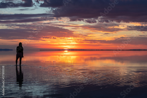 Silhouette of woman walking into the sunrise of lake Bonneville Salt Flats, Wendover, Western Utah, USA, America. Dreamy red colored clouds mirroring on the water surface creating romantic atmosphere