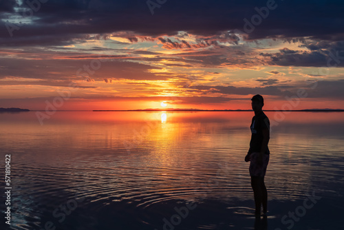 Silhouette of man walking into the sunrise of lake Bonneville Salt Flats, Wendover, Western Utah, USA, America. Dreamy red colored clouds mirroring on the water surface creating romantic atmosphere