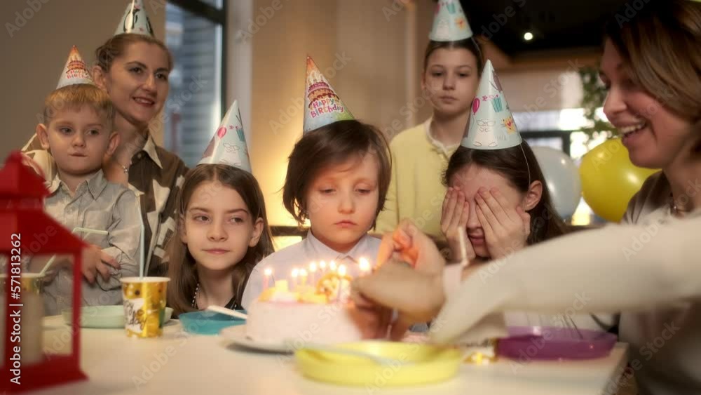 A little girl closes her eyes with her hands, makes a wish while her mother lights candles on the cake. Blows out candles on a birthday cake during a birthday celebration. Children sit at the festive A little girl closes her eyes with her hands, makes a wish while her mother lights candles on the cake. Blows out candles on a birthday cake during a birthday celebration. Children sit at the festive