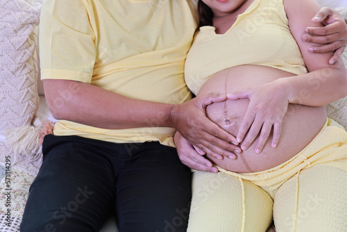 Happy asian adult couple with husband sitting and resting on sofa in living room while holding baby inside a pregnant and smile. Expectant mother preparing and waiting for baby birth during pregnancy.