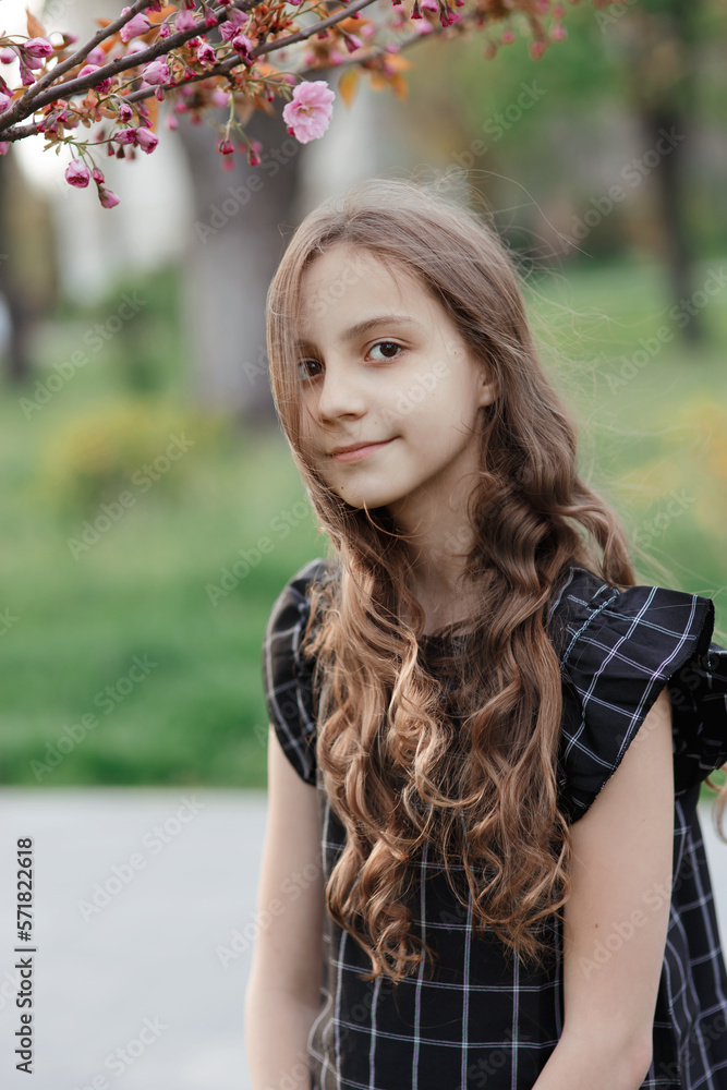 Naklejka premium Young pretty dreaming smiling girl with long hair, sakura branch with gentle pink flowers over her head, looking at camera, warm spring vertical outdoor portrait in park, purity and lightness of youth