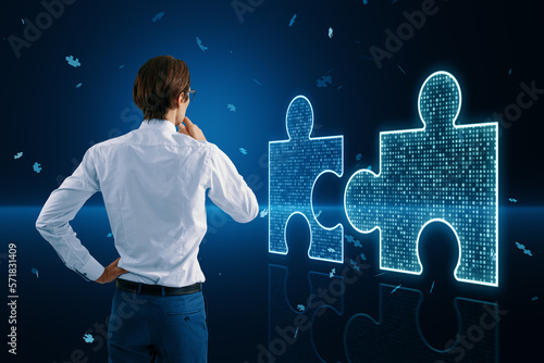 Fotografia Back view of thoughtful young businessman with glowing digital blue jigsaw puzzle hologram on dark blurry background