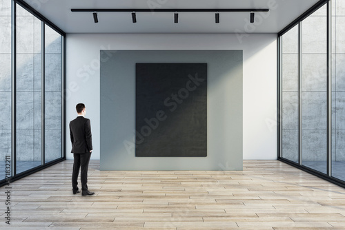Man in black suit back view looking at blank dark poster with space for your logo or text on grey partition on light background in empty exhibition hall with transparent walls and wooden floor, mockup