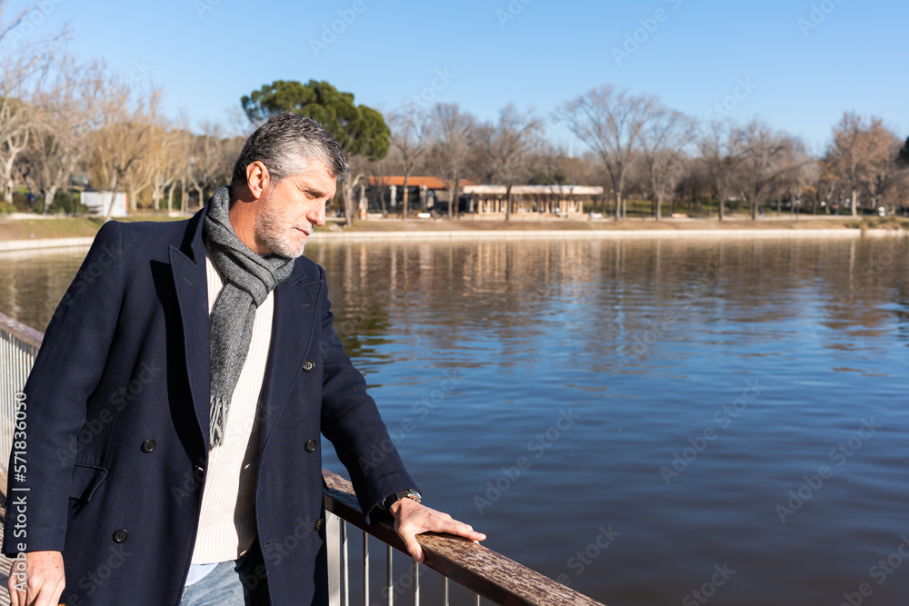 Bearded man in outerwear looking at rippling river while standing on embankment during sunny day in park Bearded man in outerwear looking at rippling river while standing on embankment during sunny day in park