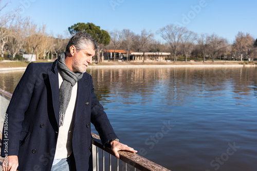 Bearded man in outerwear looking at rippling river while standing on embankment during sunny day in park