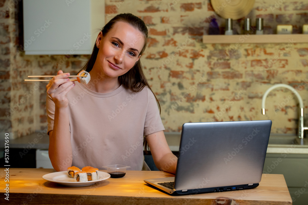 Young pretty woman sitting at her kitchen eat sushi at table home working with a laptop. Young pretty woman sitting at her kitchen eat sushi at table home working with a laptop.