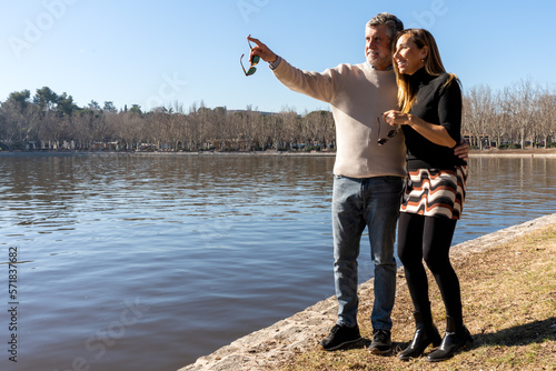 Full body of smiling gray haired man in casual outfit with sunglasses pointing at something while hugging woman on river bank near water