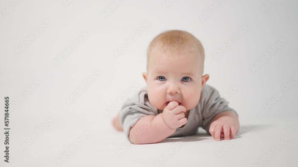 Portrait of a baby. A baby lying on his stomach looking into the camera licking his fingers, shot in close-up against a white background, looking at the cameraman Portrait of a baby. A baby lying on his stomach looking into the camera licking his fingers, shot in close-up against a white background, looking at the cameraman