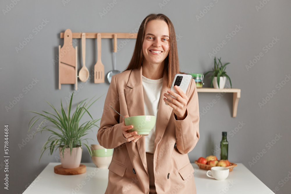 Portrait of charming cute happy young adult woman wearing beige suit standing near table on kitchen at home, eating her dish, using cell phone while having breakfast. Portrait of charming cute happy young adult woman wearing beige suit standing near table on kitchen at home, eating her dish, using cell phone while having breakfast.