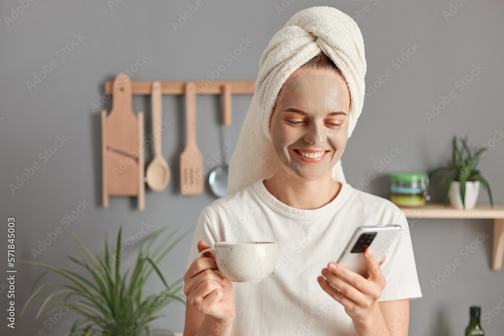 Smiling happy positive woman in a cosmetic mask and with a towel on her head posing in the kitchen, drinking coffee or tea and using smart phone, checking social networks or e-mail in morning. Smiling happy positive woman in a cosmetic mask and with a towel on her head posing in the kitchen, drinking coffee or tea and using smart phone, checking social networks or e-mail in morning.