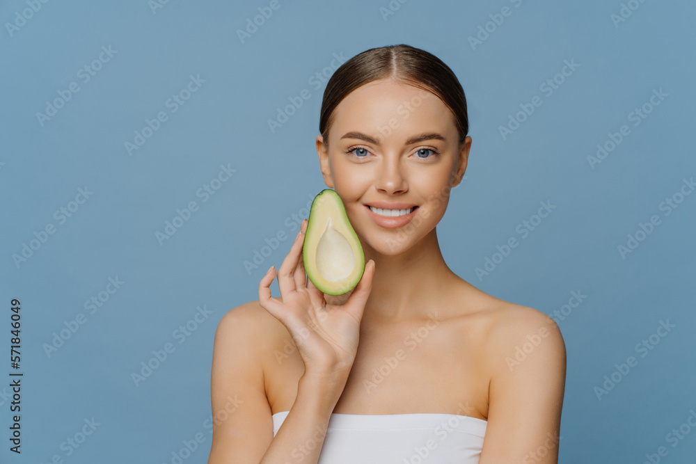 Horizontal shot of happy brunette woman holds half of avocado near face uses anti aging organic recipes stands refreshed indoor wrapped in towel isolated over blue background. Healthy diet concept Horizontal shot of happy brunette woman holds half of avocado near face uses anti aging organic recipes stands refreshed indoor wrapped in towel isolated over blue background. Healthy diet concept